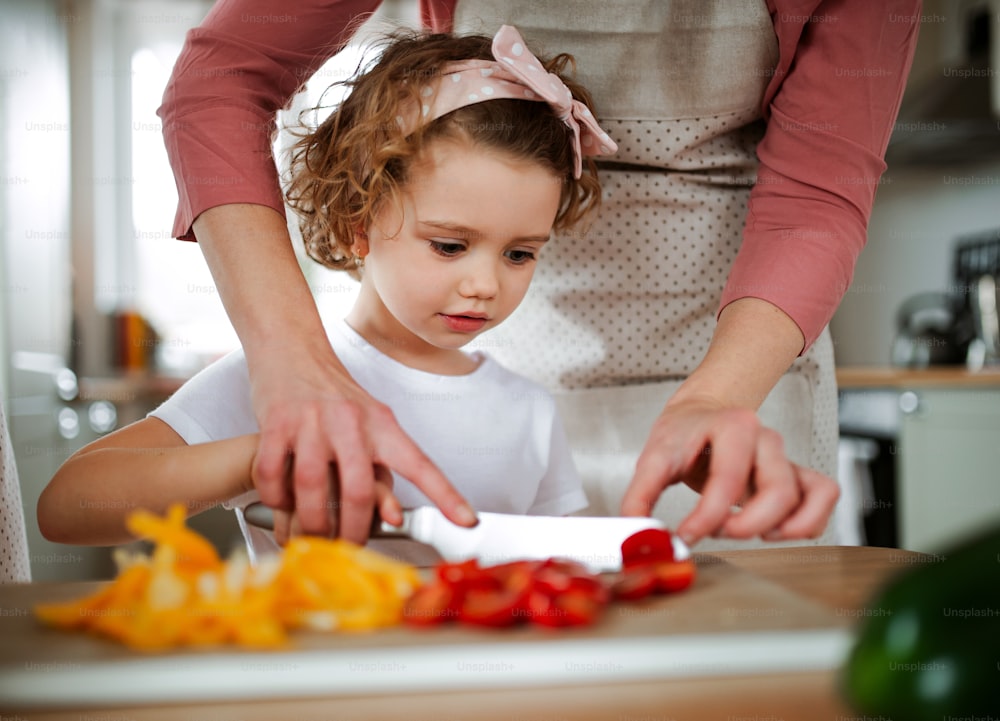 Um retrato de menina pequena com mãe irreconhecível em casa, preparando salada de legumes.