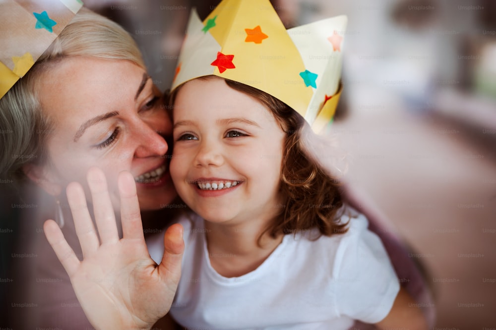 A portrait of small girl and mother with paper crown having fun at home. Shot through glass.