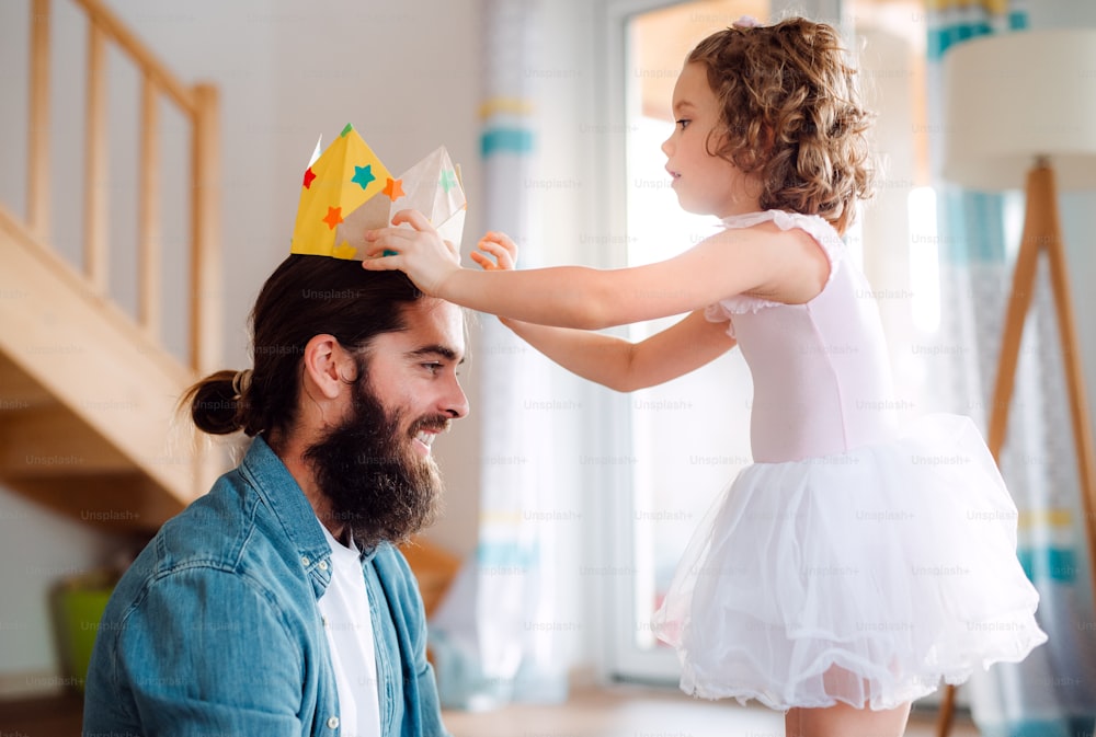 Uma vista lateral de uma menina pequena colocando uma coroa de papel na cabeça do pai em casa enquanto brincava.
