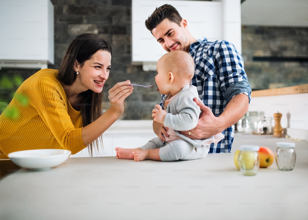 Ein Porträt einer jungen Familie, die zu Hause in einer Küche steht, eines Mannes, der ein Baby hält, und einer Frau, die es mit einem Löffel füttert.