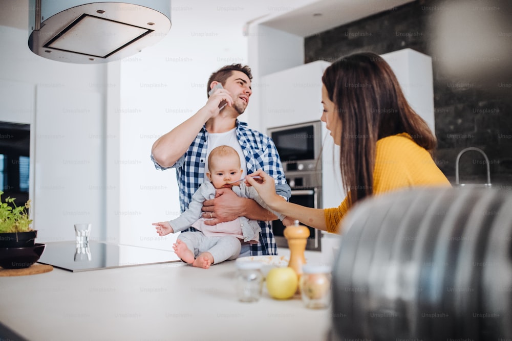 A portrait of young family standing in a kitchen at home, a man with smartphone holding a baby and a woman feeding her with a spoon.