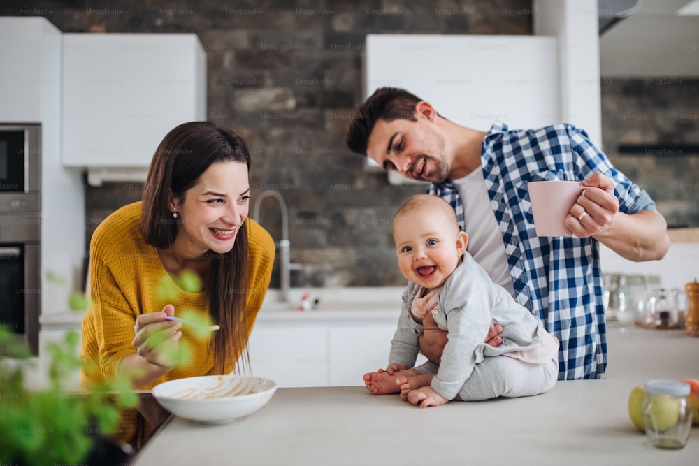 Ein Porträt einer jungen Familie, die zu Hause in einer Küche steht, eines Mannes, der ein Baby hält, und einer Frau, die es mit einem Löffel füttert.