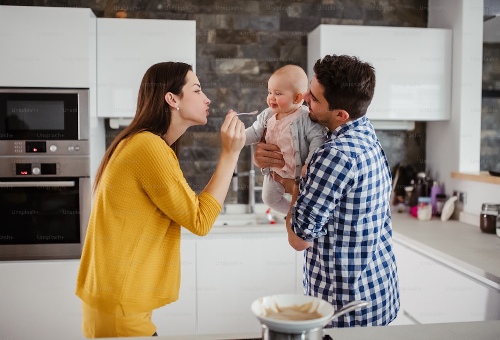 自宅の台所に立っている若い家族のポートレート、赤ん坊を抱く男性、スプーンで彼女に餌を与える女性のポートレート。