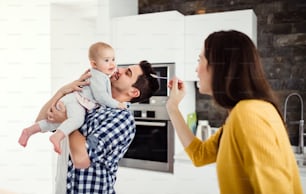 A portrait of young family standing in a kitchen at home, a man holding a baby and a woman feeding her with a spoon.