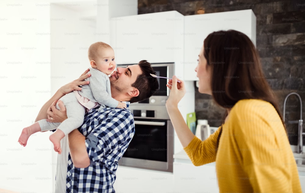 A portrait of young family standing in a kitchen at home, a man holding a baby and a woman feeding her with a spoon.