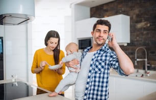 Un retrato de una familia joven de pie en una cocina en casa, un hombre haciendo una llamada telefónica y una mujer alimentando a un bebé.