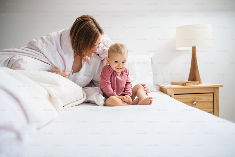 Une jeune mère heureuse avec sa petite fille assise à l’intérieur sur son lit le matin, en train de jouer.