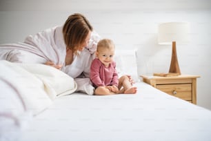 Una madre joven feliz con una hija pequeña sentada en el interior en la cama por la mañana, jugando.