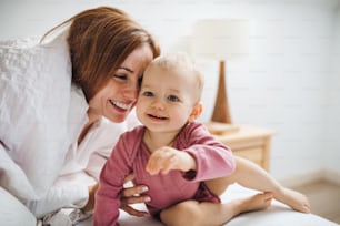 A happy young mother with little daughter sitting indoors on bed in the morning, playing.