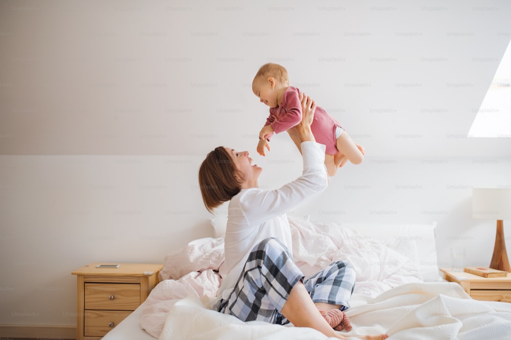 Une jeune mère heureuse avec sa petite fille assise à l’intérieur sur son lit le matin, en train de jouer.