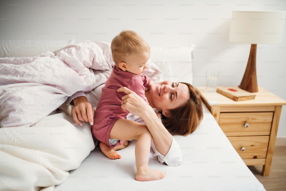 Eine glückliche junge Mutter mit kleiner Tochter, die morgens drinnen im Bett sitzt und spielt.