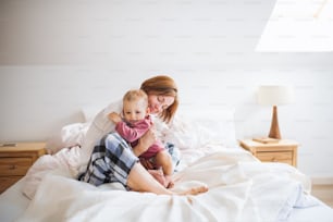 Una madre joven feliz con una hija pequeña sentada en el interior en la cama por la mañana, jugando.