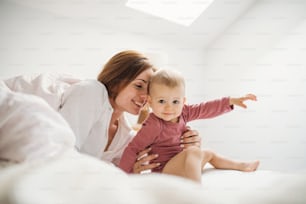 朝、小さな娘を持つ幸せな若い母親が、室内のベッドに座って遊んでいる。