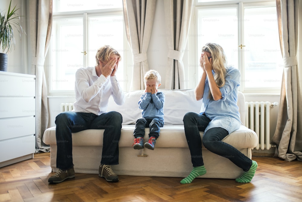 Jovens pais e filho pequeno sentados em um sofá dentro de um quarto, cobrindo os olhos ao jogar peekaboo.