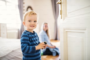 A cheerful toddler boy with young mother in the background inside in a bedroom.