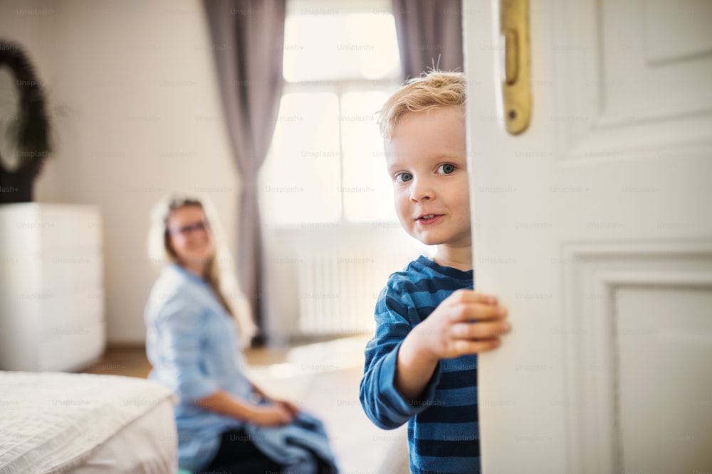 A toddler boy with unrecognizable young mother in the background inside in a bedroom, holding door.