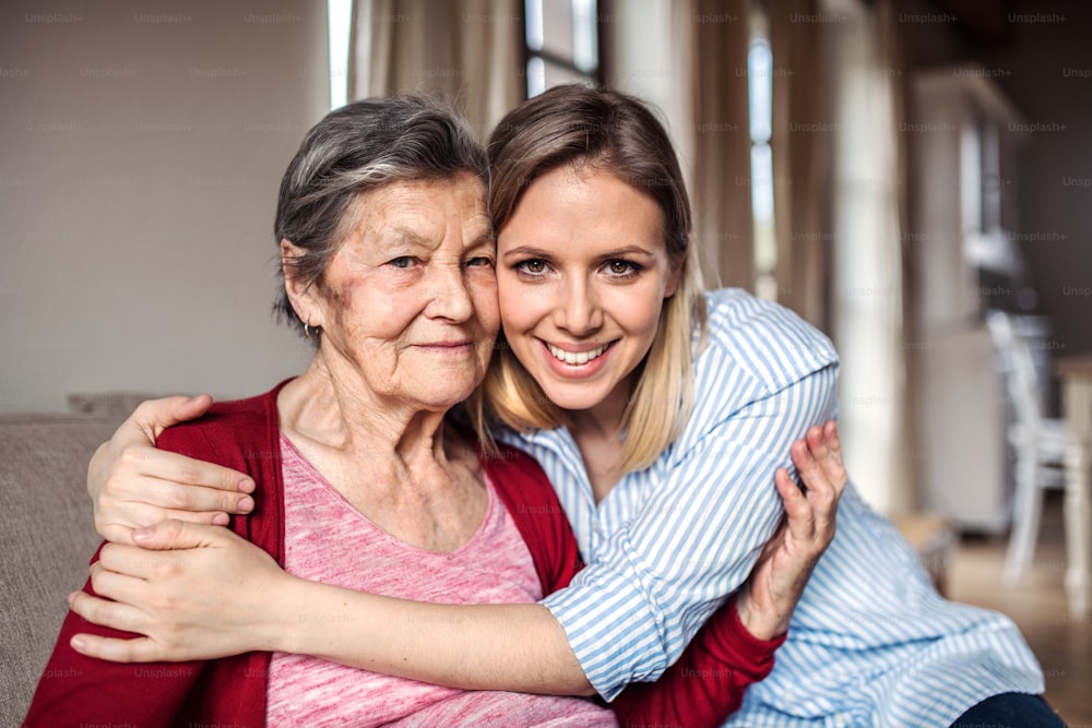 Retrato de uma avó idosa com uma neta adulta sentada no sofá de casa, abraçada.
