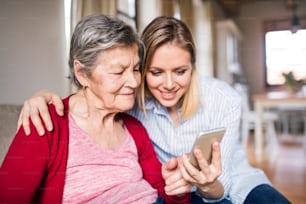 An elderly grandmother and adult granddaughter with smartphone at home.