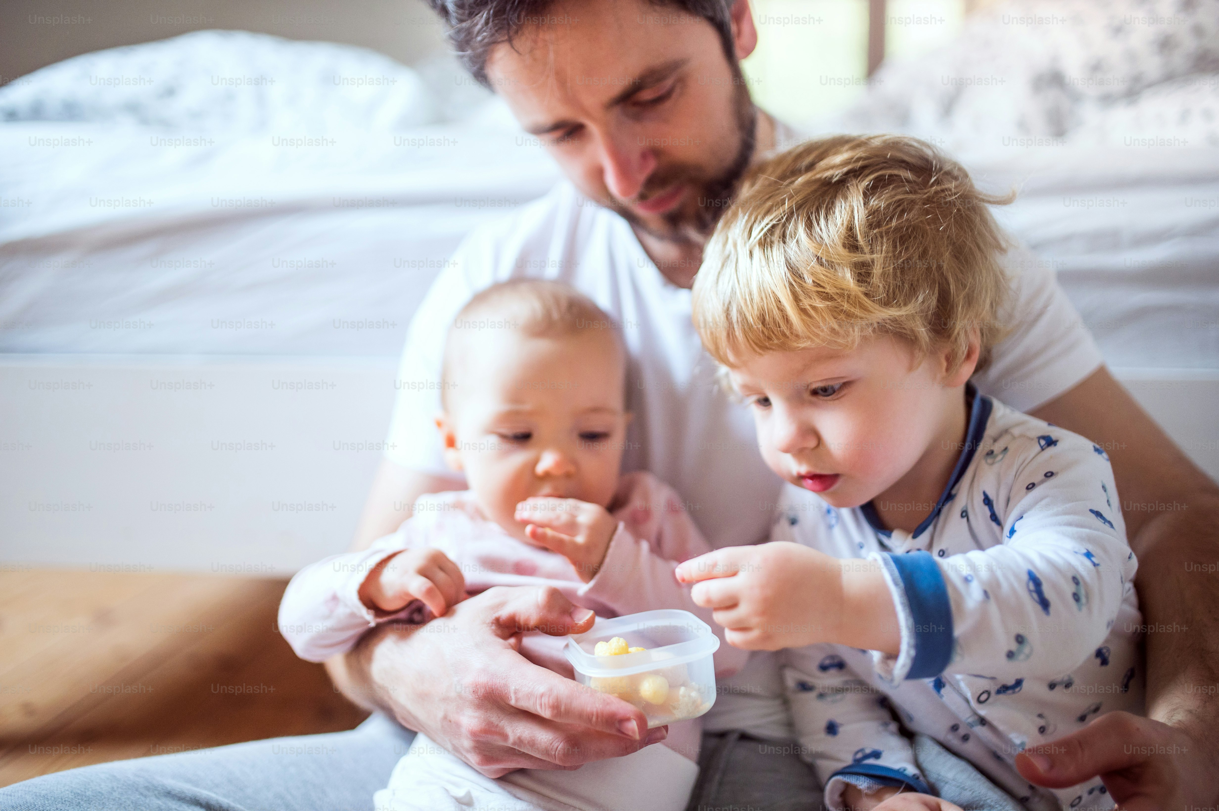 Hübscher Vater mit Kleinkindern, die Fingerfood im Schlafzimmer zu Hause essen. Vaterschaftsurlaub.