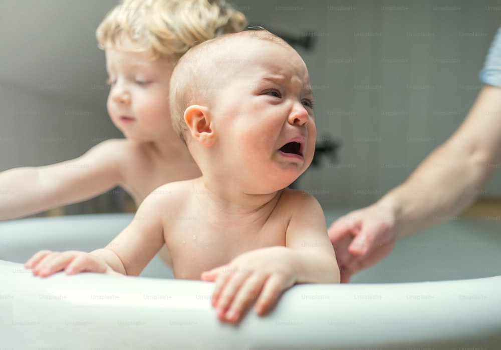 An unrecognizable father washing two unhappy toddlers in the bath in the bathroom at home. A small girl crying. Paternity leave.