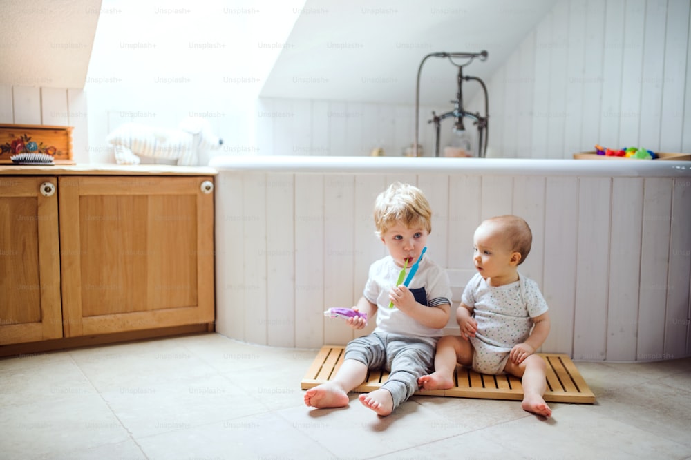 Two toddler children sitting on the floor and brushing teeth in the bathroom at home.