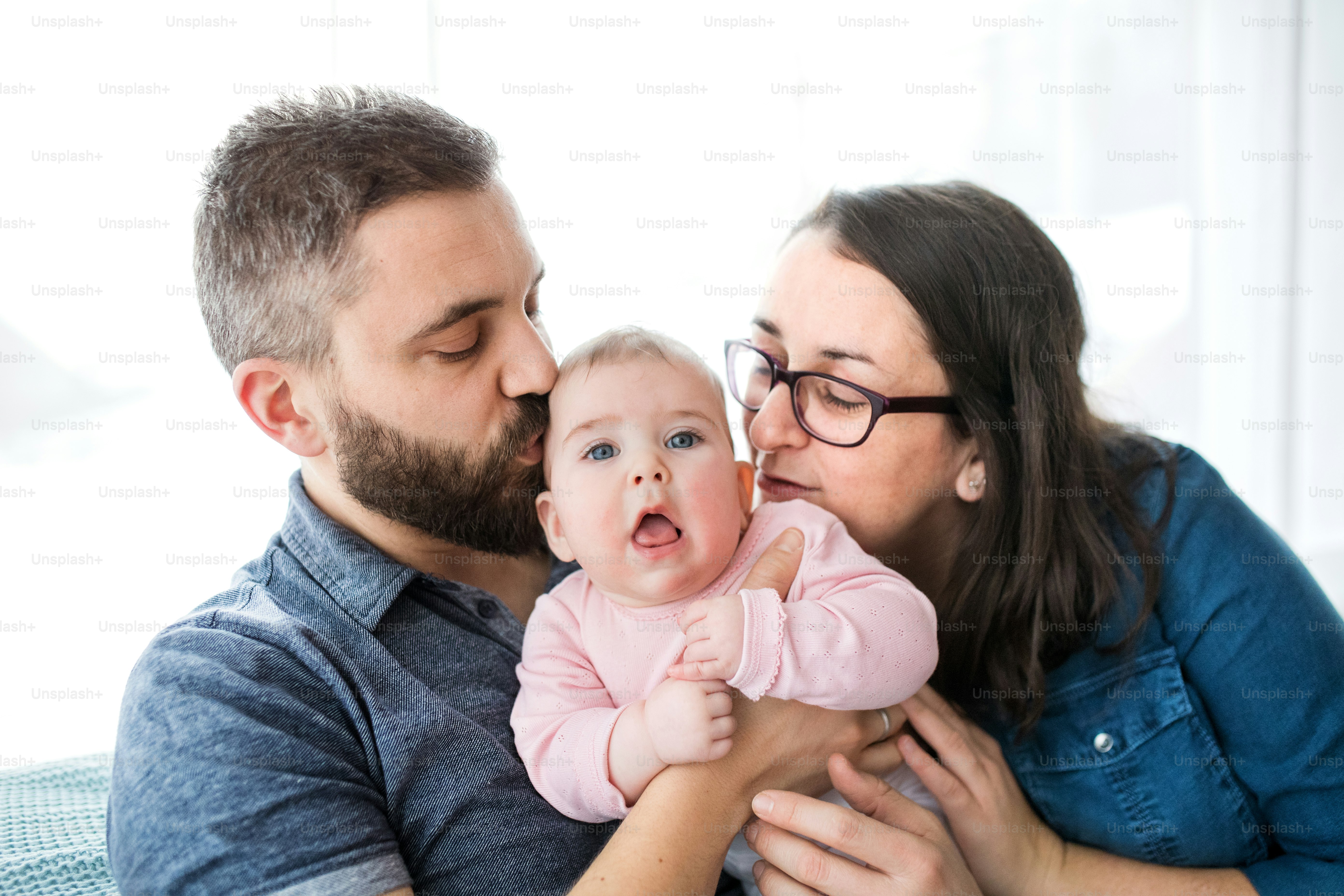 A portrait of young family with baby daughter sitting indoors on a sofa, kissing her.