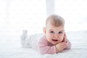 A portrait of unhappy baby girl lying on her tummy on bed indoors.