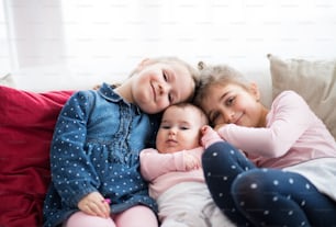 Ein Porträt von drei glücklichen kleinen Mädchen, die drinnen auf einem Sofa sitzen.