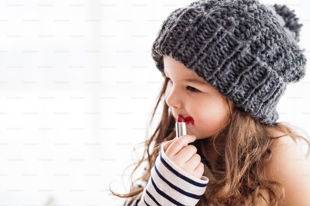 A small happy girl in striped T-shirt at home, wearing a hat and applying a lipstick. Copy space.