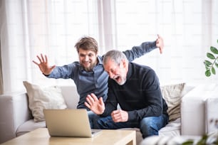 Hijo hipster y su padre mayor con computadora portátil en casa, divirtiéndose. Dos generaciones en interior.