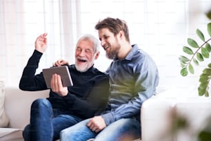 Hipster son and his senior father with tablet at home. Two generations indoors, having fun.Hipster son and his senior father at home. Two generations indoors, having fun.Hipster son with his senior father at home. Two generations indoors, having fun.