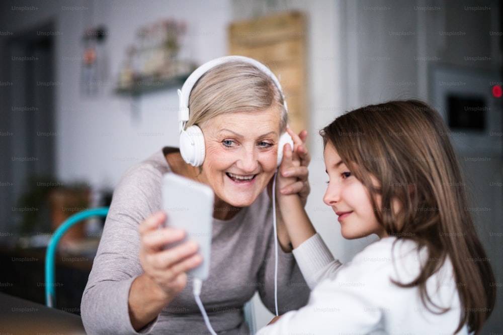 小さな女の子と祖母が家にスマートフォンとヘッドフォンを持っています。家族と世代のコンセプト。