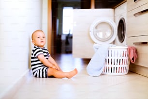 Lindo bebé junto a la lavadora mashine en la cocina. Cesta de lavandería en el piso.