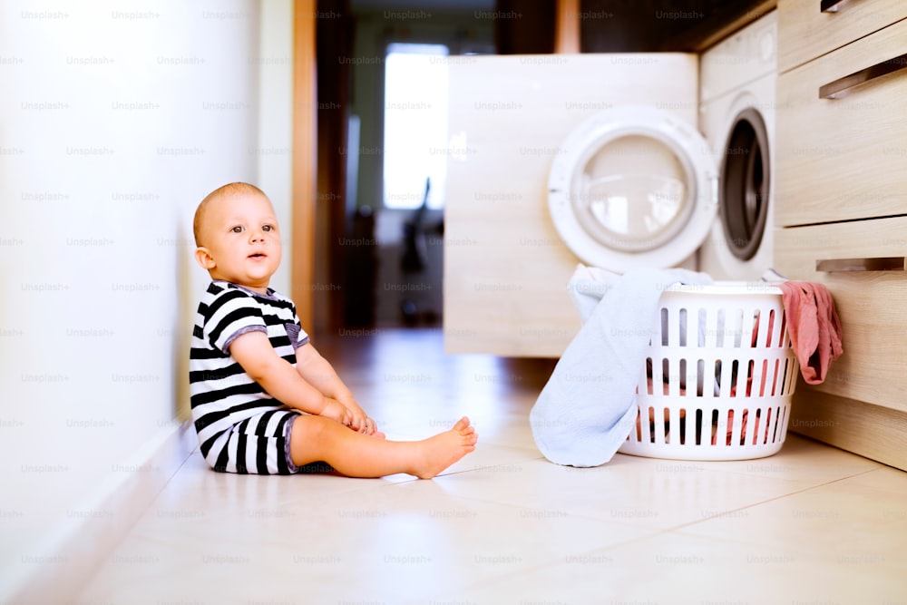 Cute baby boy by the washing mashine in the kitchen. Laundry basket on the floor.