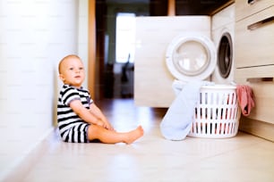 Cute baby boy by the washing mashine in the kitchen. Laundry basket on the floor.