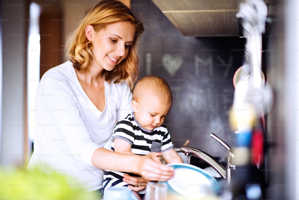 Mãe jovem com um filho bebê fazendo tarefas domésticas. Mulher bonita e menino lavando os pratos.