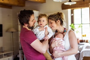 Beautiful young parents at home standing in the kitchen holding their cute little children in the arms.