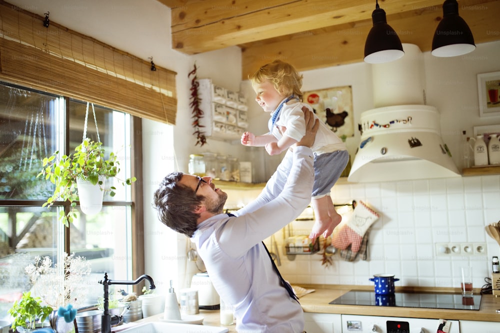 Young businessman coming home from work, in the kitchen with his little son holding him high in the air.