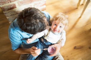 床に座ってかわいい幼い息子にヨーグルトを食べさせる家の若い父親。