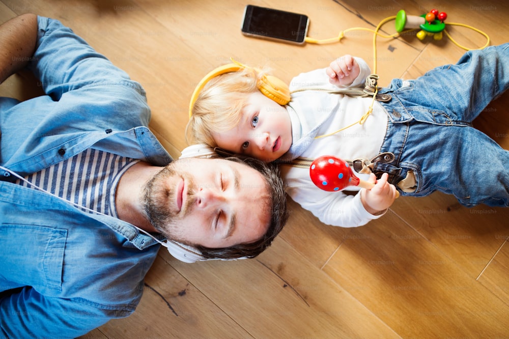Junger Vater und sein kleiner Sohn mit Smartphone und Kopfhörern, zu Hause Musik hörend, auf Holzboden liegend, Spielzeugmusikinstrumente um sie herum.