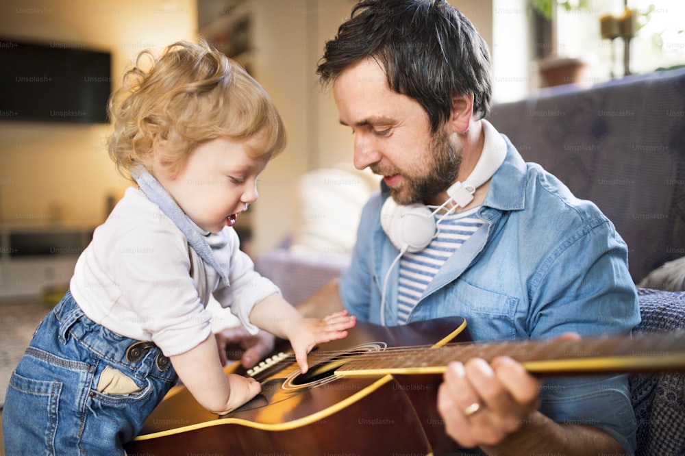 Young father at home sitting on wooden floor, his cute little son playing guitar that he is holding.
