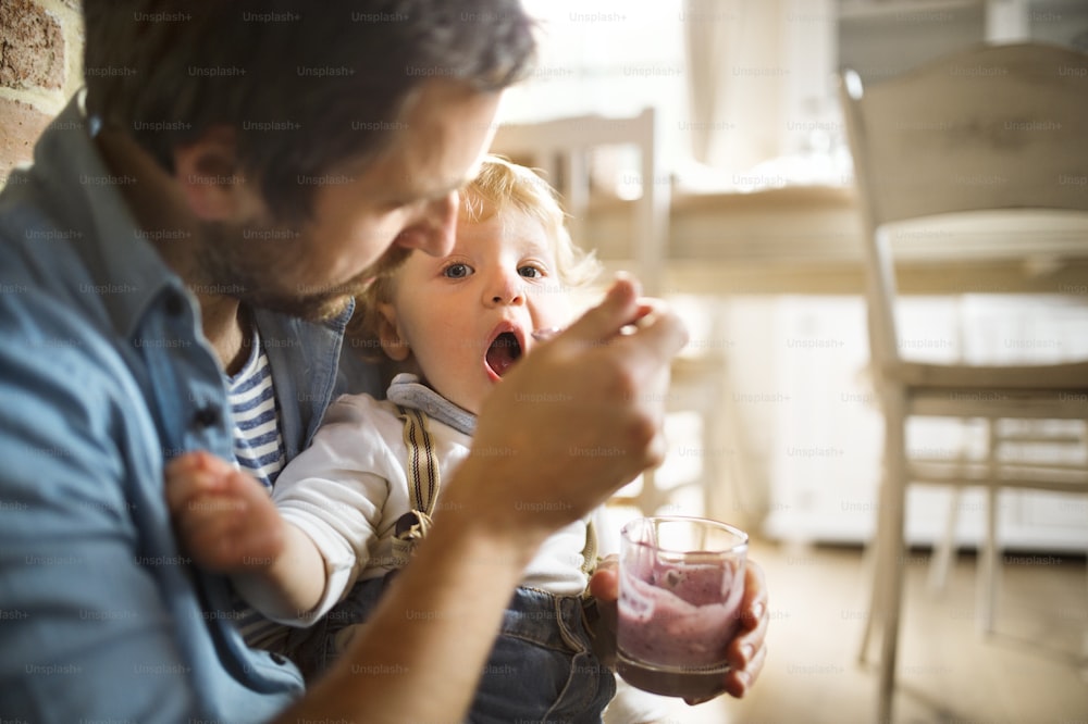 Junger Vater zu Hause sitzt auf dem Boden und füttert seinen süßen kleinen Sohn mit Joghurt.