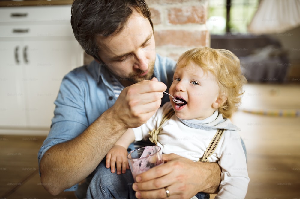 Junger Vater zu Hause sitzt auf dem Boden und füttert seinen süßen kleinen Sohn mit Joghurt.