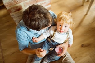 Padre joven en casa sentado en el suelo alimentando a su lindo hijo pequeño con yogur.