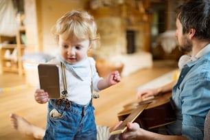 Young father at home sitting on wooden floor, playing guitar, his cute little son holding smart phone, playing with it.