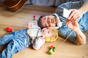 Young father and his little son with smart phone and earphones, listening music at home, lying on wooden floor, musical toys surrounding them.