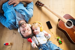 Junger Vater und sein kleiner Sohn mit Smartphone und Kopfhörern, zu Hause Musik hörend, auf Holzboden liegend, Gitarre und andere Musikinstrumente um sie herum.