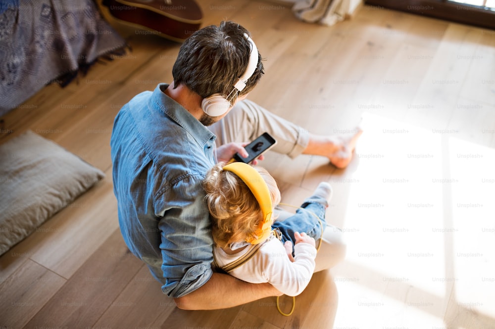 Padre joven y su hijo pequeño con teléfono inteligente y auriculares, escuchando música en casa, sentados en el piso de madera. Vista trasera.