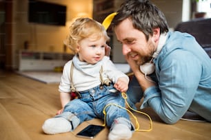 Junger Vater und sein kleiner Sohn mit Smartphone und Kopfhörern, zu Hause Musik hörend, auf Holzboden sitzend.