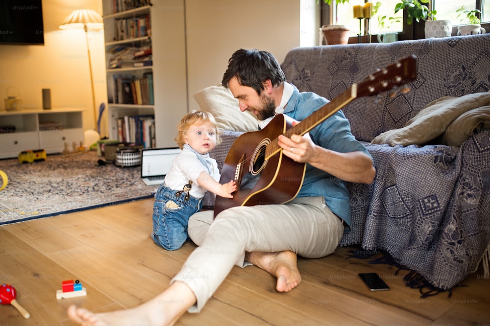 Young father at home sitting on wooden floor, his cute little son playing guitar that he is holding.